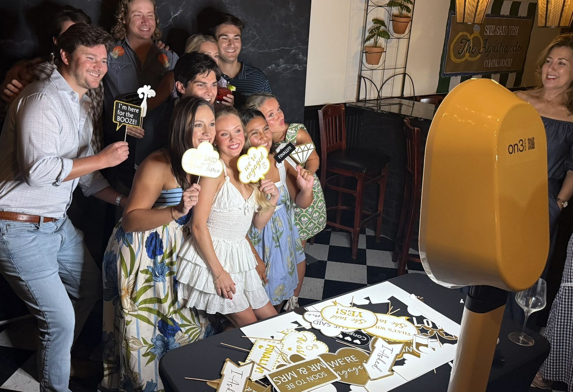 A group of smiling guests pose with fun photo props in front of a yellow On3 Media digital photo booth at an engagement party. The scene includes a table full of props and a festive sign in the background reading “She Said Yes! The Aguillards Coming Soon.” The setting is lively and joyful, capturing the energy of the celebration.
